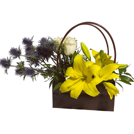 Floral arrangement with yellow lilies, white roses, and purple thistles in a brown basket on a white background.